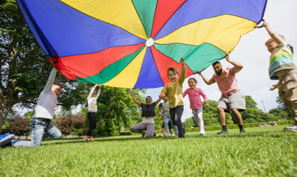 Kinder spielen mit einem bunten Fallschirm und ein Junge rennt darunter hindurch.