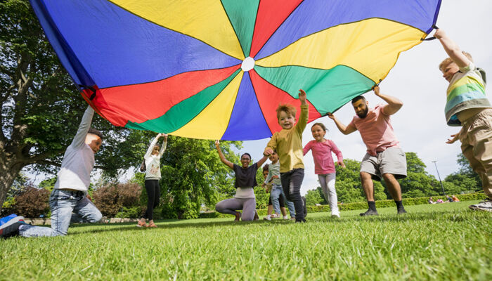 Kinder spielen mit einem bunten Fallschirm und ein Junge rennt darunter hindurch.