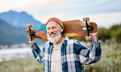 An older man in a plaid shirt is grinning as he carries his colorful skateboard over his shoulder, with headphones in his ears
