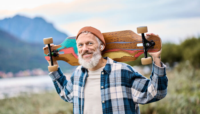 Älterer Mann im karierten Hemd trägt grinsend sein buntes Skateboard im Nacken und hat Kopfhörer in den Ohren