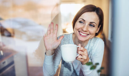 Eine junge Frau mit einer Tasse schaut aus dem Fenster und winkt zum Abschied.
