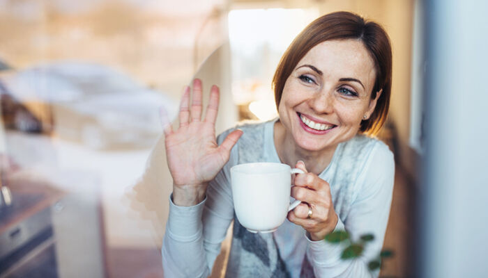 Eine junge Frau mit einer Tasse schaut aus dem Fenster und winkt zum Abschied.