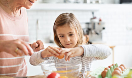 An older woman watches as a young girl cracks an egg into a bowl