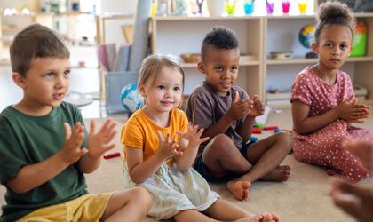 Eine Gruppe kleiner Kindergartenkinder sitzt drinnen im Klassenzimmer auf dem Boden und klatscht.