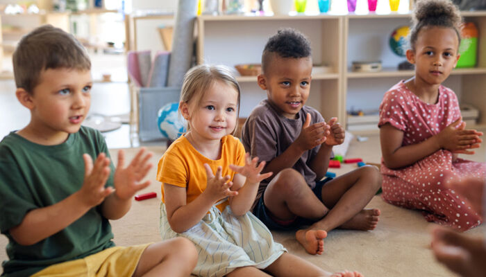 Eine Gruppe kleiner Kindergartenkinder sitzt drinnen im Klassenzimmer auf dem Boden und klatscht.