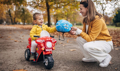 Frau hockt vor kleinem Jungen auf einem Fahrzeug und reicht ihm einen Helm