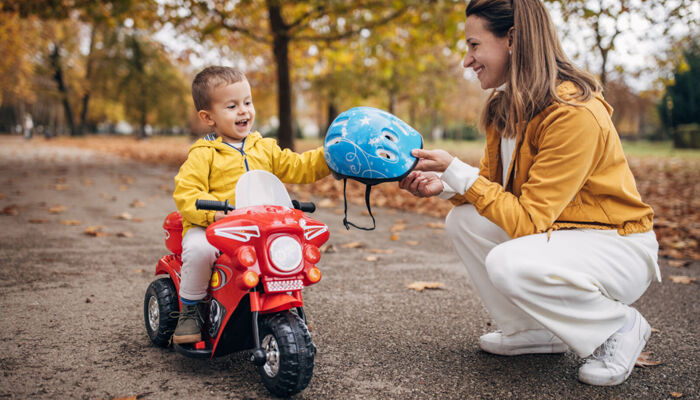 Frau hockt vor kleinem Jungen auf einem Fahrzeug und reicht ihm einen Helm