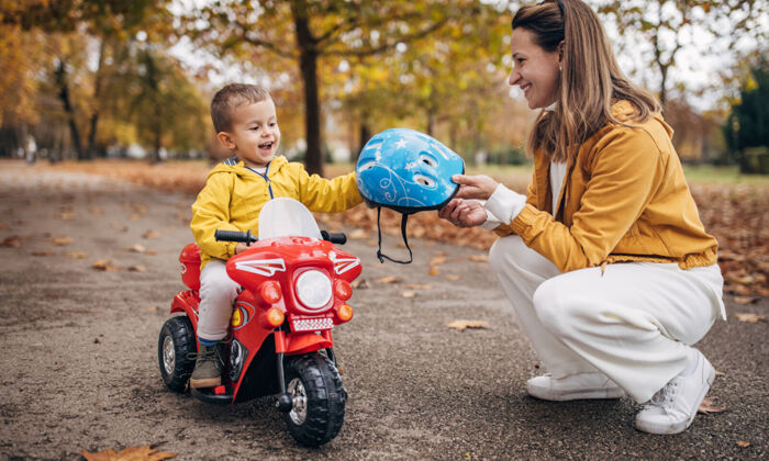 Frau hockt vor kleinem Jungen auf einem Fahrzeug und reicht ihm einen Helm