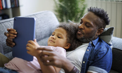 Ein glücklicher afroamerikanischer Vater und seine Tochter nutzen ein Tablet auf dem Sofa liegend