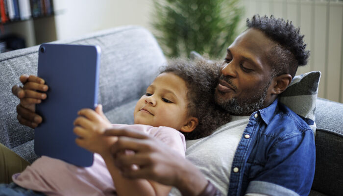 Ein glücklicher afroamerikanischer Vater und seine Tochter nutzen ein Tablet auf dem Sofa liegend